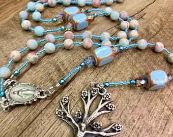 May include: A rosary with light blue, peach, and tan beads. The rosary features light blue square beads with brown accents. The silver crucifix is in the shape of a tree with a figure of Jesus. A silver medallion is also present.