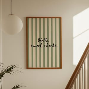 Puede incluir: Un estampado enmarcado con un borde marrón presenta rayas verticales crema y verde claro. El texto "Hello sweet cheeks" está escrito en cursiva. El estampado cuelga de una pared blanca cerca de una escalera y una planta.