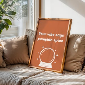 Peut inclure: Une affiche encadrée avec un fond orange et le texte "Your vibe says pumpkin spice" en blanc. Un dessin de boule de cristal blanche est en dessous du texte. Le cadre est de couleur marron clair. L'affiche est posée contre un canapé.