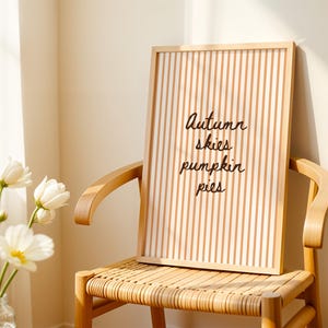 Peut inclure: Une œuvre d'art encadrée avec un motif à rayures verticales et les mots "Autumn skies pumpkin pies" en écriture noire. L'œuvre est posée sur une chaise en bois avec une assise tressée, à côté d'un vase de fleurs blanches.