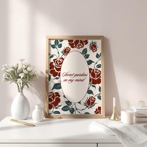 Peut inclure: Une affiche encadrée avec un motif floral de roses bordeaux et de feuilles vertes sur fond crème. L'affiche présente un ovale central avec le texte "Secret gardens in my mind". Le cadre est en bois.