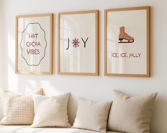 Puede incluir: Tres impresiones artísticas enmarcadas con temática invernal. Una impresión dice "Hot Cocoa Vibes", otra dice "JOY" con un copo de nieve, y la última muestra un patín de hielo y dice "Ice, Ice, Jolly". Los marcos son de color marrón claro.