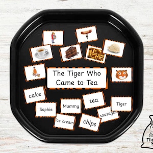 May include: A black tray with orange and white cards featuring illustrations and words related to the book "The Tiger Who Came to Tea". The cards include words like "cake", "tea", "chips", "sausages", "ice cream", "tiger", "mummy", and "Sophie".