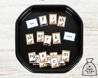 May include: A black tray with educational cards for spelling. The cards spell out "fish" and "shark", with individual letters and illustrations of a fish and a snail. The Story Sack logo is in the bottom right corner.
