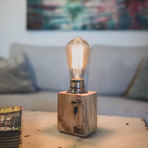 Puede incluir: Una lámpara de madera con una bombilla y una cerradura. La lámpara está sobre una mesa de madera con una revista en la mesa. La lámpara está hecha por "The Clamp".
