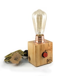Puede incluir: Una lámpara de mesa de madera con una base de cobre y una bombilla de estilo vintage. La lámpara tiene un botón rojo en el lateral y un cable amarillo y verde.