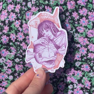 May include: A sticker featuring a pink and purple illustration of a woman with long hair holding a large fish. The woman is wearing a dress and has a smaller fish on her head. The sticker has a white background and is on a pink and green floral fabric.