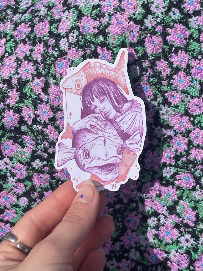 May include: A sticker featuring a pink and purple illustration of a woman with long hair holding a large fish. The woman is wearing a dress and has a smaller fish on her head. The sticker has a white background and is on a pink and green floral fabric.