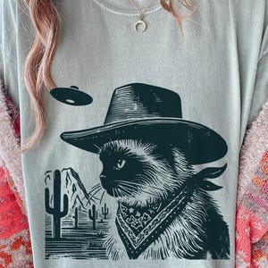 Puede incluir: Camiseta verde claro con un gráfico en blanco y negro de un gato con sombrero de vaquero y pañuelo, con un OVNI encima. El diseño incluye un paisaje desértico con cactus y montañas.