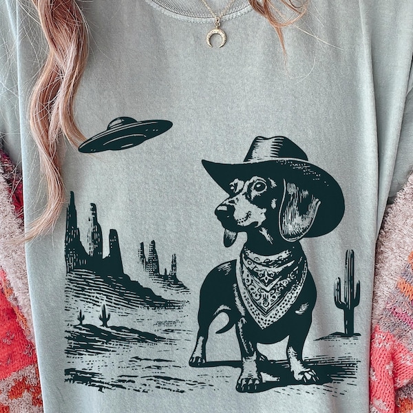 Könnte beinhalten: Hellgraues T-Shirt mit einer schwarz-weißen Grafik eines Dackels mit Cowboyhut und Halstuch, mit einem UFO über einer Wüstenlandschaft. Das Shirt hat einen Rundhalsausschnitt und kurze Ärmel.