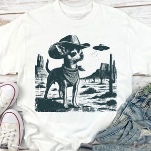 May include: White t-shirt featuring a black and white graphic of a Chihuahua wearing a cowboy hat and bandana, set against a desert landscape with a UFO. The design includes cacti and mountains, creating a unique and whimsical scene.