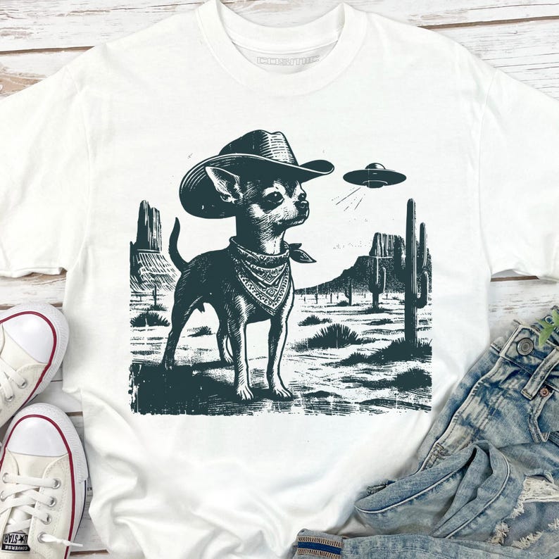 May include: White t-shirt featuring a black and white graphic of a Chihuahua wearing a cowboy hat and bandana, set against a desert landscape with a UFO. The design includes cacti and mountains, creating a unique and whimsical scene.