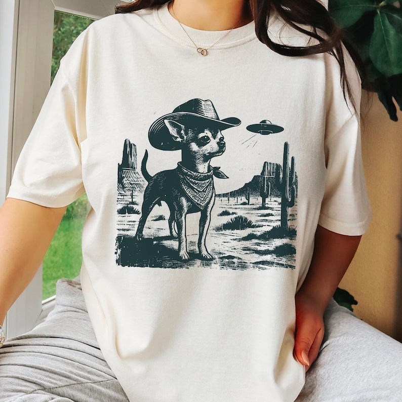 May include: Cream-colored t-shirt featuring a graphic of a chihuahua wearing a cowboy hat and bandana, set against a desert landscape with a UFO. The design is in dark green, creating a unique and playful aesthetic.