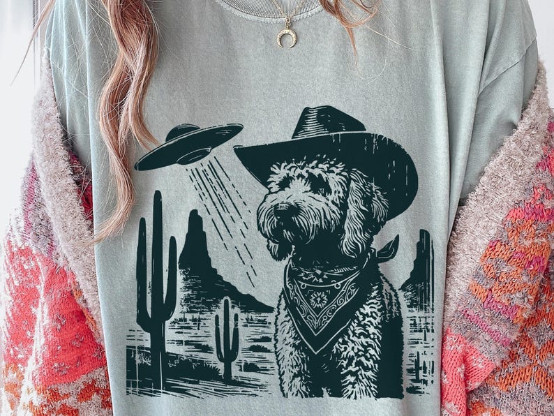 K&ouml;nnte beinhalten: Hellgraues T-Shirt mit einem schwarz-wei&szlig;en Aufdruck eines Hundes mit Cowboyhut und Halstuch, der von einem UFO angestrahlt wird. Der Hintergrund zeigt Kakteen und W&uuml;stenberge.