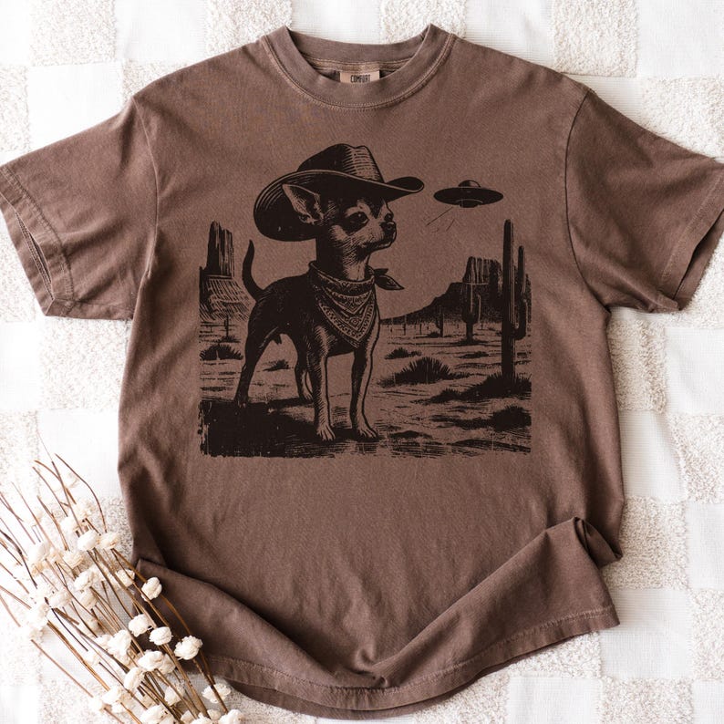 May include: Brown t-shirt featuring a graphic of a chihuahua wearing a cowboy hat and bandana, set against a desert landscape with a UFO. The design is in a vintage style.