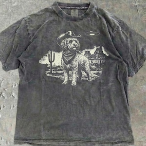 Peut inclure: Un t-shirt gris anthracite délavé à l'acide avec une impression graphique représentant un chien portant un chapeau de cowboy et un bandana. Le chien se tient dans un paysage désertique avec un cactus, des rochers et une soucoupe volante.