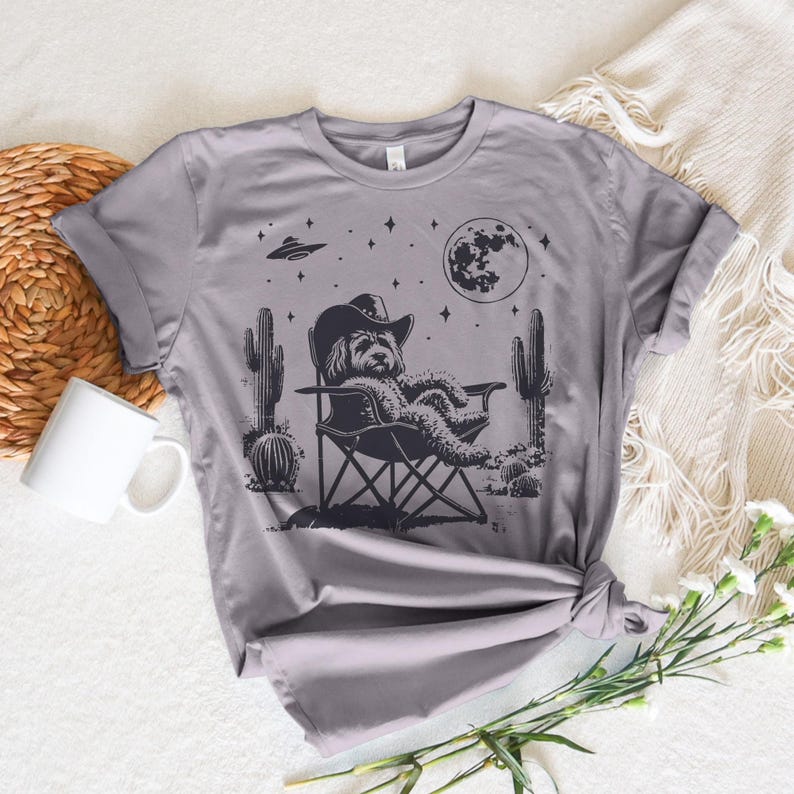 Op de afbeelding: Een grijs T-shirt met een zwart-witte illustratie van een hond die een cowboyhoed draagt en op een campingstoel zit onder een halve maan en sterren. De hond is omringd door cactussen.