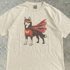 May include: Cream-colored t-shirt featuring a graphic of a dog dressed as a superhero. The dog wears a red cape and a red and gold emblem. The design is printed on a short-sleeved, crew neck t-shirt.