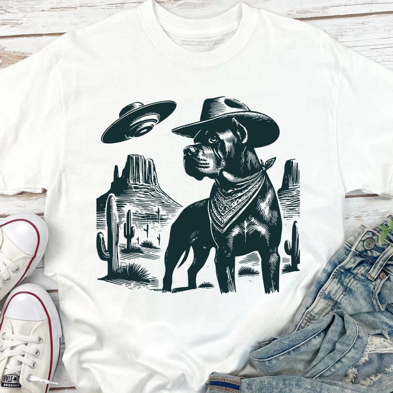 May include: White t-shirt featuring a black graphic of a dog wearing a cowboy hat and bandana, set against a desert landscape with cacti and a UFO. The design is a humorous blend of Western and sci-fi themes.