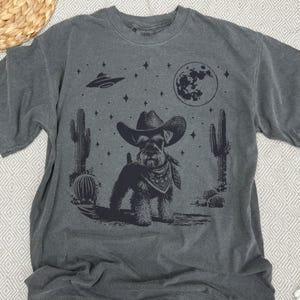 May include: Dark gray t-shirt featuring a black graphic of a dog wearing a cowboy hat and bandana, set against a desert landscape with cacti, a full moon, stars, and a UFO. The shirt has a vintage, worn look.