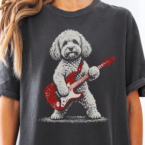 May include: A charcoal gray short-sleeve t-shirt featuring a graphic print of a fluffy, light-colored dog standing on its hind legs while playing a red electric guitar. The dog has a textured, curly coat and is positioned in the center of the shirt.