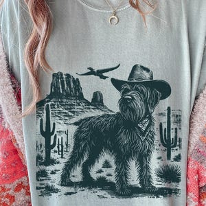 Puede incluir: Camiseta azul claro con una ilustración en blanco y negro de un perro con sombrero de vaquero y pañuelo, ambientada en un paisaje desértico con cactus y montañas. Un pájaro vuela por encima.