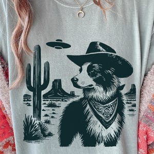 Puede incluir: Camiseta gris claro con un gráfico en blanco y negro de un perro con sombrero de vaquero y pañuelo, ambientado en un paisaje desértico con un cactus y un OVNI. El perro es el foco principal del diseño.