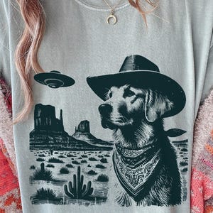 May include: A light gray t-shirt featuring a dog wearing a cowboy hat and bandana, with a desert landscape and a UFO in the background. The graphic is in black and white.