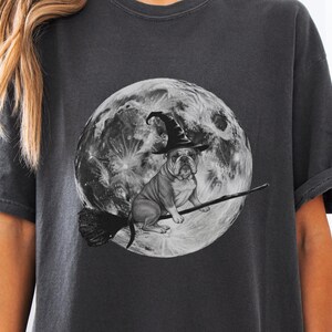 May include: Dark gray t-shirt featuring a bulldog wearing a witch hat and riding a broomstick in front of a full moon. The graphic is in black and white, creating a Halloween theme.