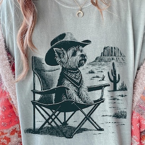 Pu&ograve; includere: T-shirt azzurra con un'illustrazione verde scuro di un cane con cappello da cowboy e bandana, seduto su una sedia da campeggio. Lo sfondo mostra un paesaggio desertico con un cactus e formazioni rocciose.
