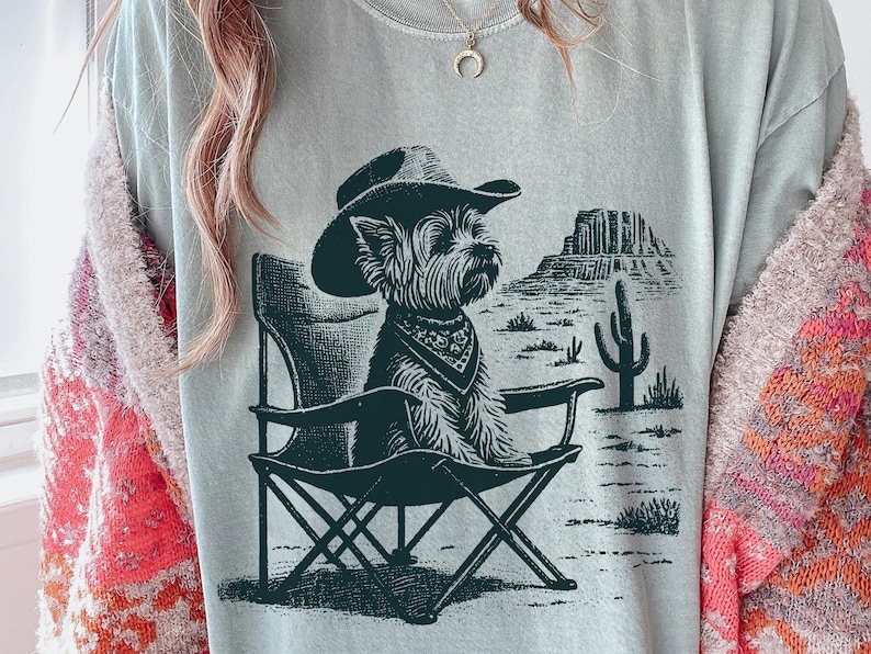 Pu&ograve; includere: T-shirt azzurra con un'illustrazione verde scuro di un cane con cappello da cowboy e bandana, seduto su una sedia da campeggio. Lo sfondo mostra un paesaggio desertico con un cactus e formazioni rocciose.
