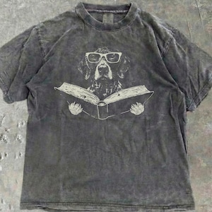 May include: A charcoal grey short-sleeve t-shirt with a vintage-style acid wash finish. The center graphic features a line-art illustration of a dog wearing thick-rimmed glasses while holding an open book with its front paws.