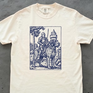 Puede incluir: Camiseta color crema con una ilustración azul marino de un caballero a caballo, sobre un perro. El caballero está blindado y sostiene una espada. El perro lleva una corona. El fondo incluye un castillo y árboles.
