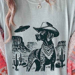 May include: A light blue t-shirt featuring a graphic of a dog wearing a cowboy hat and bandana, with a UFO hovering above a desert landscape. The design is in black and white.
