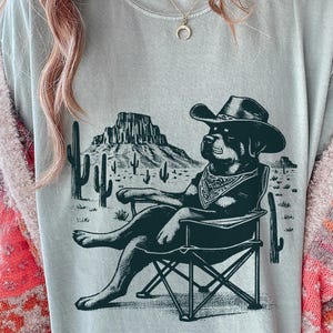 Könnte beinhalten: Hellblaues T-Shirt mit einem schwarz-weißen Grafikdruck eines Hundes mit Cowboyhut und Halstuch, der in einem Klappstuhl entspannt. Der Hintergrund zeigt eine Wüstenlandschaft mit Kakteen und Bergen.
