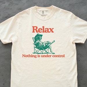 Pode incluir: Camiseta creme com a palavra "Relax" em letras vermelhas acima de uma ilustração verde de um cachorro com chapéu de cowboy relaxando em uma cadeira dobrável. Abaixo da imagem, o texto diz "Nothing is under control".