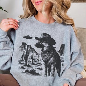 Peut inclure: Un sweat-shirt gris clair avec un motif noir et blanc. Le motif représente un chien portant un chapeau de cow-boy et un bandana, sur fond de paysage désertique avec des cactus et un OVNI.