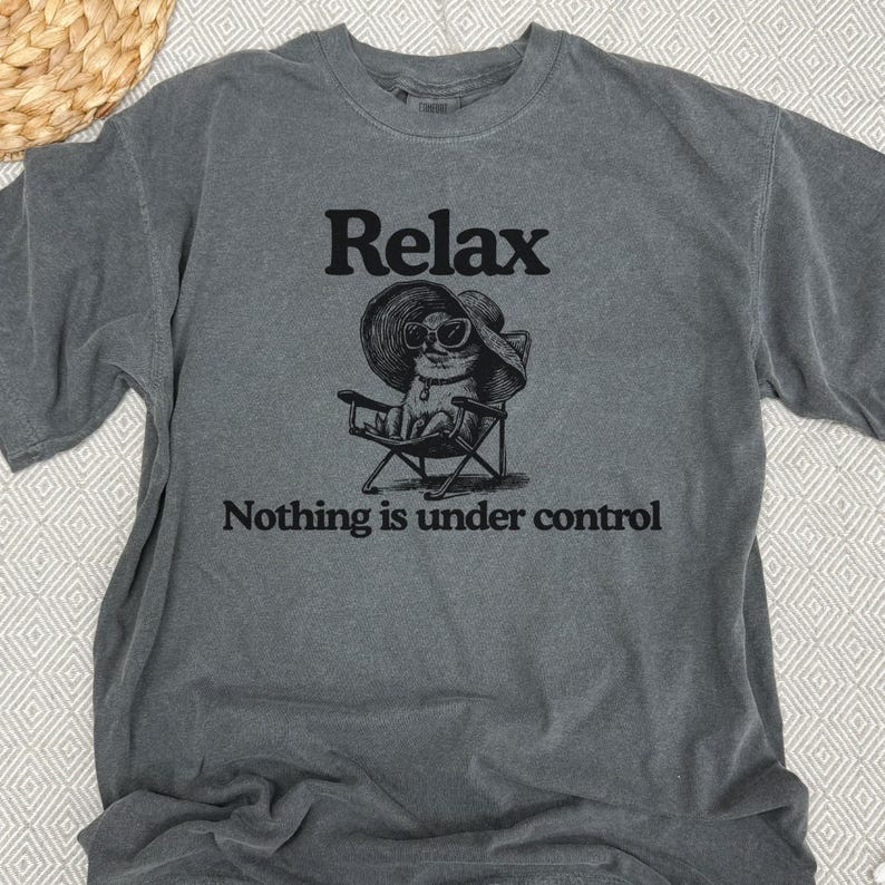 May include: Gray t-shirt with the word "Relax" above a graphic of a cat wearing sunglasses and a sun hat, seated in a folding chair. The text "Nothing is under control" is below the graphic.