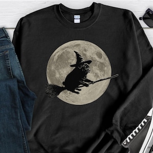 May include: Black sweatshirt with a graphic of a pug in a witch hat flying on a broomstick against a full moon. The design is in black and white. The sweatshirt is shown with blue jeans and black sunglasses.