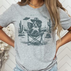 May include: A light gray t-shirt featuring a graphic of a dog wearing a cowboy hat, seated in a folding chair in a desert scene with cacti and a UFO. The dog is the central focus of the design.