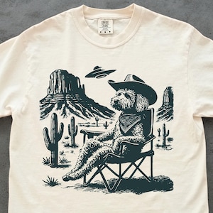 May include: Cream-colored t-shirt featuring a desert scene with a dog wearing a cowboy hat and bandana, seated in a folding chair. The graphic includes cacti, rock formations, and a UFO. The shirt has a crew neck and short sleeves.
