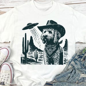 K&ouml;nnte beinhalten: Wei&szlig;es T-Shirt mit einem schwarz-wei&szlig;en Grafikdesign. Es zeigt einen Hund mit Cowboyhut und Halstuch, der von einem UFO angestrahlt wird. Das Design beinhaltet Kakteen und Berge in einer W&uuml;stenlandschaft.