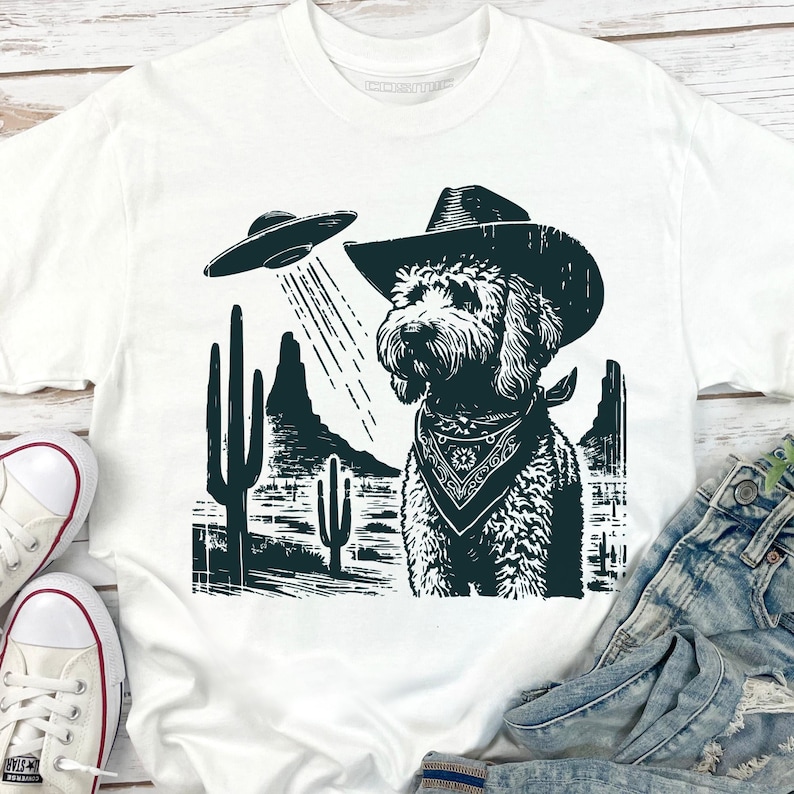 K&ouml;nnte beinhalten: Wei&szlig;es T-Shirt mit einem schwarz-wei&szlig;en Grafikdesign. Es zeigt einen Hund mit Cowboyhut und Halstuch, der von einem UFO angestrahlt wird. Das Design beinhaltet Kakteen und Berge in einer W&uuml;stenlandschaft.