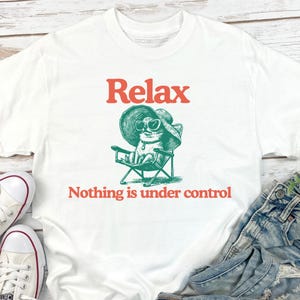 May include: White t-shirt with the word "Relax" in red above a green illustration of a cat wearing sunglasses and a hat, sitting in a beach chair. The text "Nothing is under control" is below the illustration.