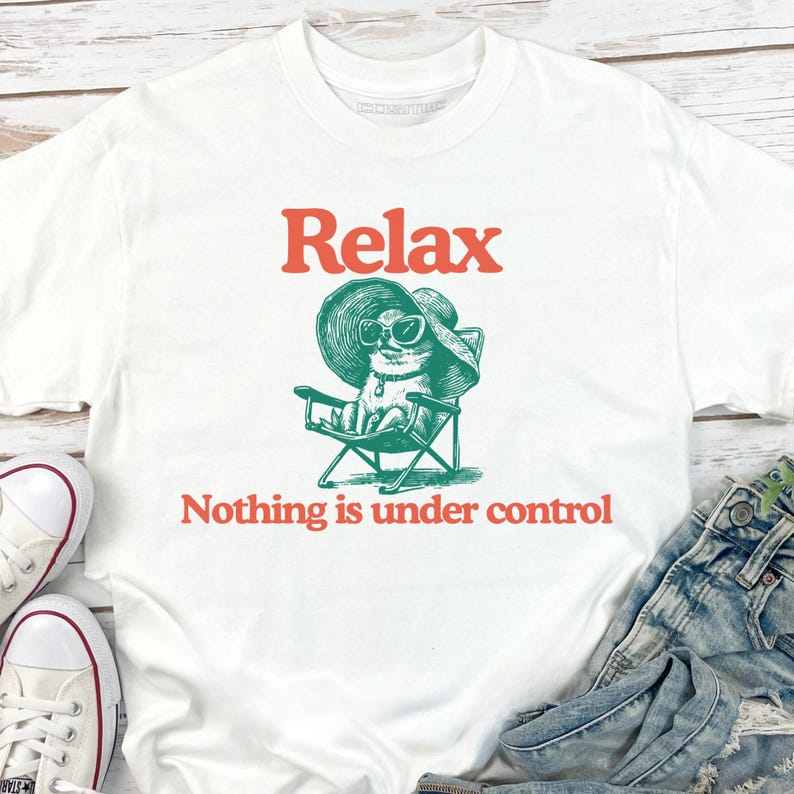 May include: White t-shirt with the word "Relax" in red above a green illustration of a cat wearing sunglasses and a hat, sitting in a beach chair. The text "Nothing is under control" is below the illustration.