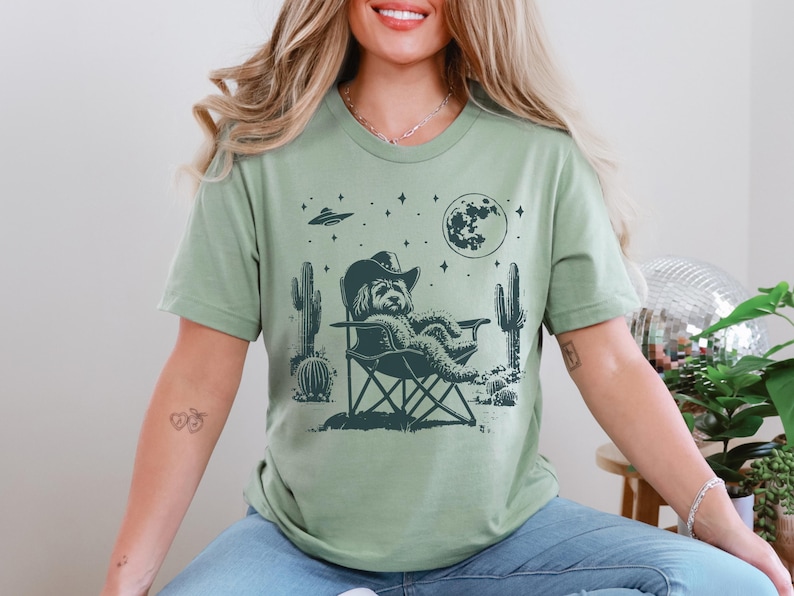 Op de afbeelding: Een groen T-shirt met een afbeelding van een hond die een cowboyhoed draagt en in een campingstoel zit onder een halve maan. De hond wordt omringd door cactussen en sterren.