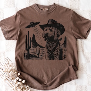K&ouml;nnte beinhalten: Braunes T-Shirt mit einem schwarzen Aufdruck eines Hundes mit Cowboyhut und Halstuch, vor einer W&uuml;stenkulisse mit einem UFO. Das Design konzentriert sich auf den Hund, mit der W&uuml;stenlandschaft und dem UFO im Hintergrund.