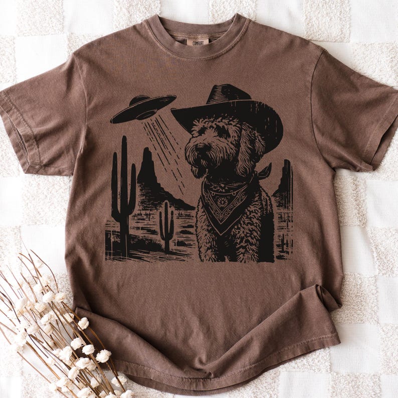 K&ouml;nnte beinhalten: Braunes T-Shirt mit einem schwarzen Aufdruck eines Hundes mit Cowboyhut und Halstuch, vor einer W&uuml;stenkulisse mit einem UFO. Das Design konzentriert sich auf den Hund, mit der W&uuml;stenlandschaft und dem UFO im Hintergrund.