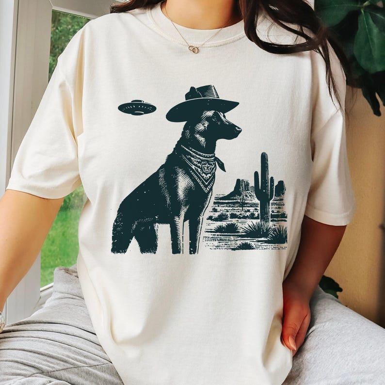 May include: Cream-colored t-shirt featuring a black graphic of a dog wearing a cowboy hat and bandana, with a desert landscape and UFO. The dog is the central focus, with the desert scene and flying saucer adding a unique touch.