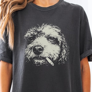 Peut inclure: Un t-shirt à manches courtes gris anthracite avec une impression graphique représentant un chien aux poils bouclés tenant une cigarette dans la gueule. L'illustration du chien est en couleur crème claire, contrastant avec le tissu sombre.